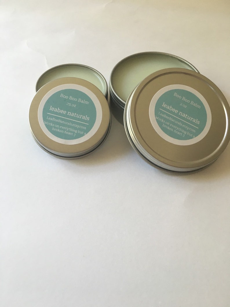 May include: Several small, round, silver tins of "Boo Boo Balm" are displayed. Each tin has a light blue label with the product name, size, and the "leabee naturals" brand name. The text on the label also includes a website address and a tagline.