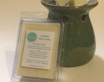 Op de afbeelding: Een transparante plastic verpakking met een wax melt met het label "LeaBee Wax Melts Persian Lilac". Het label beschrijft de geur als een natuurlijke mix van bijenwas en sojawas. De wax melt ligt op een groene keramische wax warmer met twee witte wax melts erop.