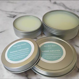May include: Four silver tins of balm, each with a light green, creamy texture. Two tins have labels that read "Boo Boo Balm" and "leabee naturals". The labels are light blue with white text. The balm is in a variety of sizes.