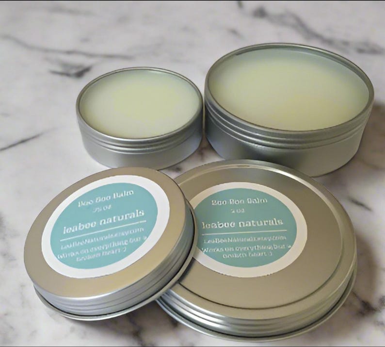 May include: Four silver tins of balm, each with a light green, creamy texture. Two tins have labels that read "Boo Boo Balm" and "leabee naturals". The labels are light blue with white text. The balm is in a variety of sizes.