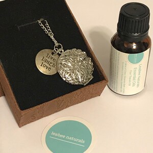Puede incluir: Un collar de plata con un colgante de medallón que dice "live laugh love" y una pequeña botella de aceite esencial con la etiqueta "LeaBee Naturals Essential Oils Four Thieves Oil" y una pegatina que dice "leabee naturals".