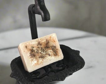 May include: A black cast iron soap dish with a decorative design and a faucet-shaped stand. The soap dish has a white soap bar with dried flowers and gold glitter inside.