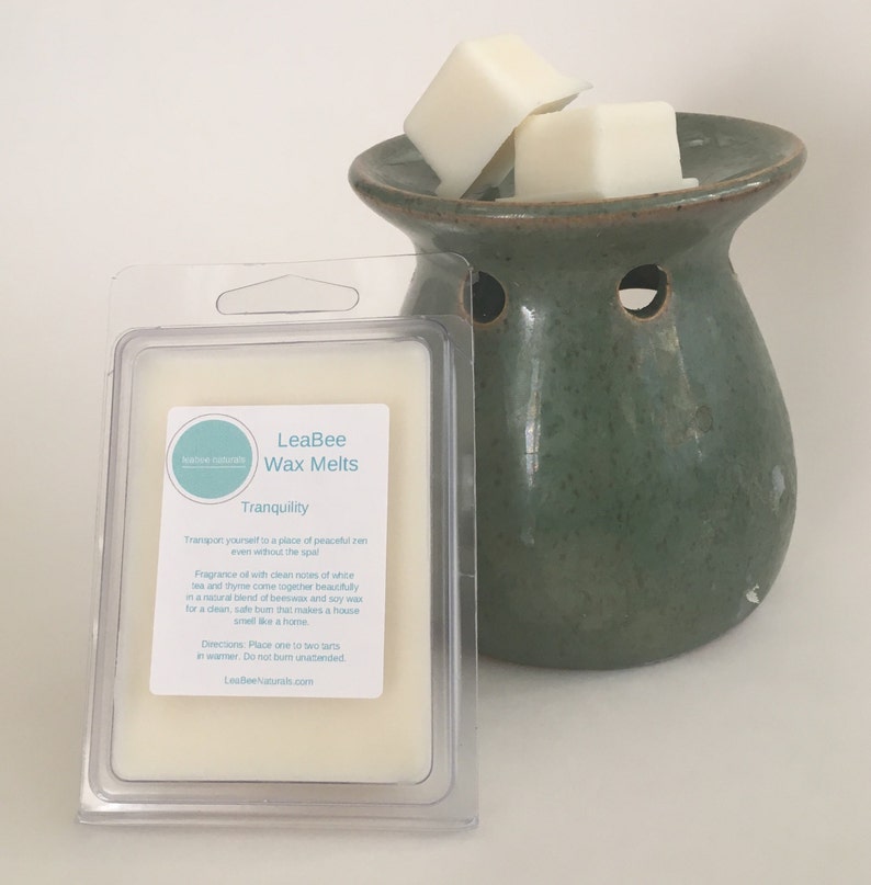 Op de afbeelding: Een transparante plastic verpakking met een witte was smelt met het label "LeaBee Wax Melts Tranquility". De was smelt zit in een witte keramische schaal met een groene keramische waswarmer.