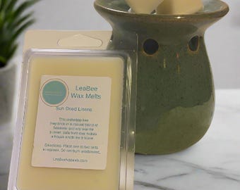 Op de afbeelding: Een doorzichtige plastic container met een wax melt erin. De wax melt is lichtgeel van kleur en heeft een label met de tekst "LeaBee Wax Melts Sun Dried Linens". Het label bevat ook een beschrijving van de geur. De container staat op een wit marmeren oppervlak. Op de achtergrond is een groene keramische wax warmer te zien met twee wax melts erin.