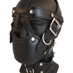 May include: Black leather head harness with multiple straps, buckles, and metal hardware. The harness covers the head and mouth, with a chin strap and a strap across the forehead. The product is designed for restraint.