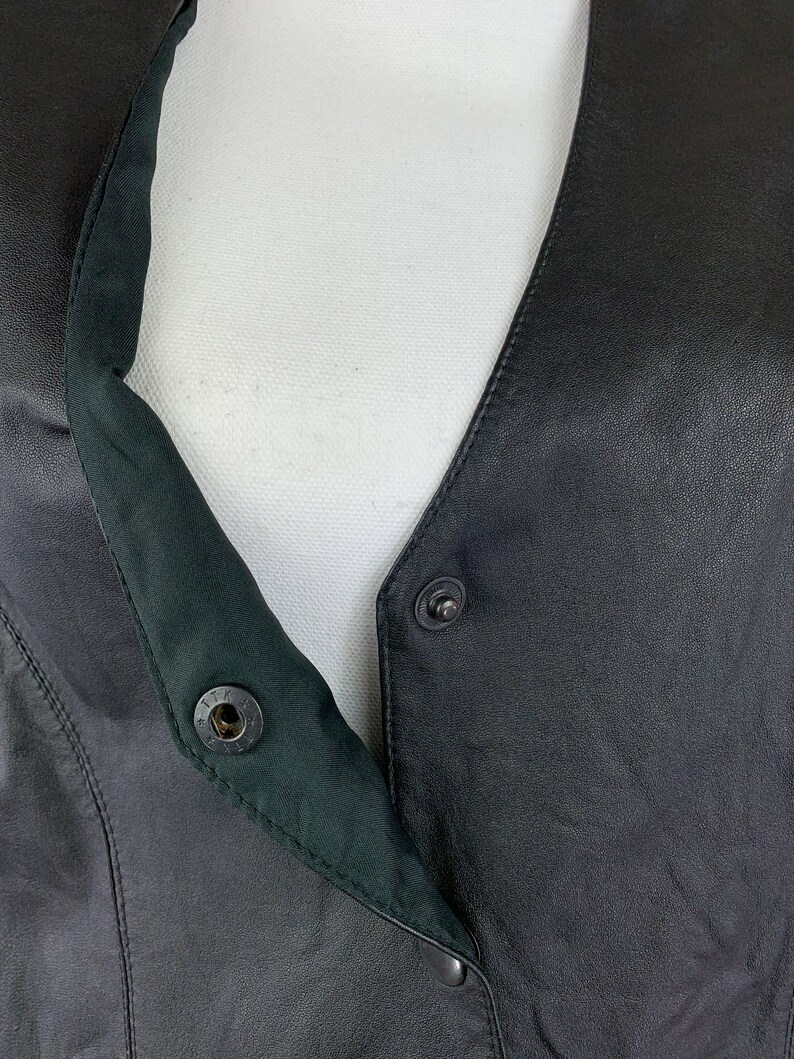 May include: Black leather vest with a snap closure and a green lining. The vest has a V-neck and a button closure at the top.