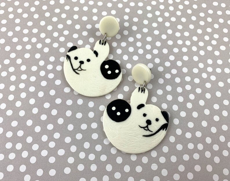 May include: A pair of white dangle earrings with black panda bear designs. The panda bears are holding up their paws and have black eyes and noses.