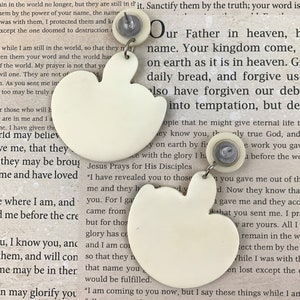 May include: A pair of white ceramic earrings shaped like apples. The earrings have a small silver post and back. The earrings are hanging from a white background with text about Jesus.