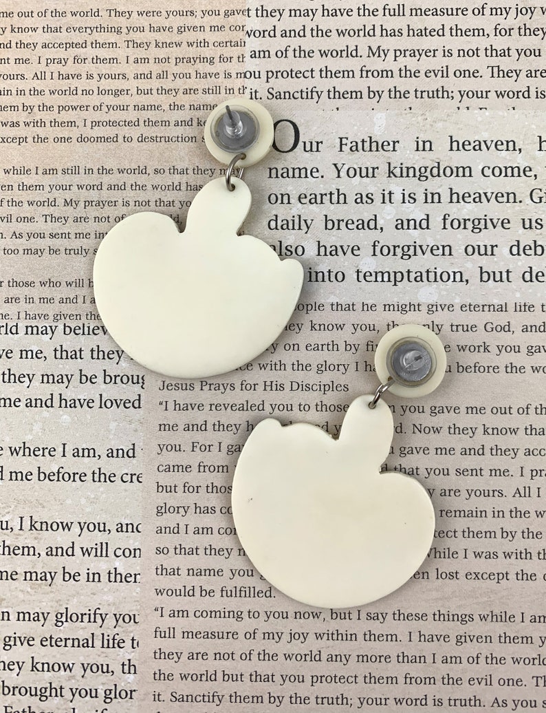 May include: A pair of white ceramic earrings shaped like apples. The earrings have a small silver post and back. The earrings are hanging from a white background with text about Jesus.