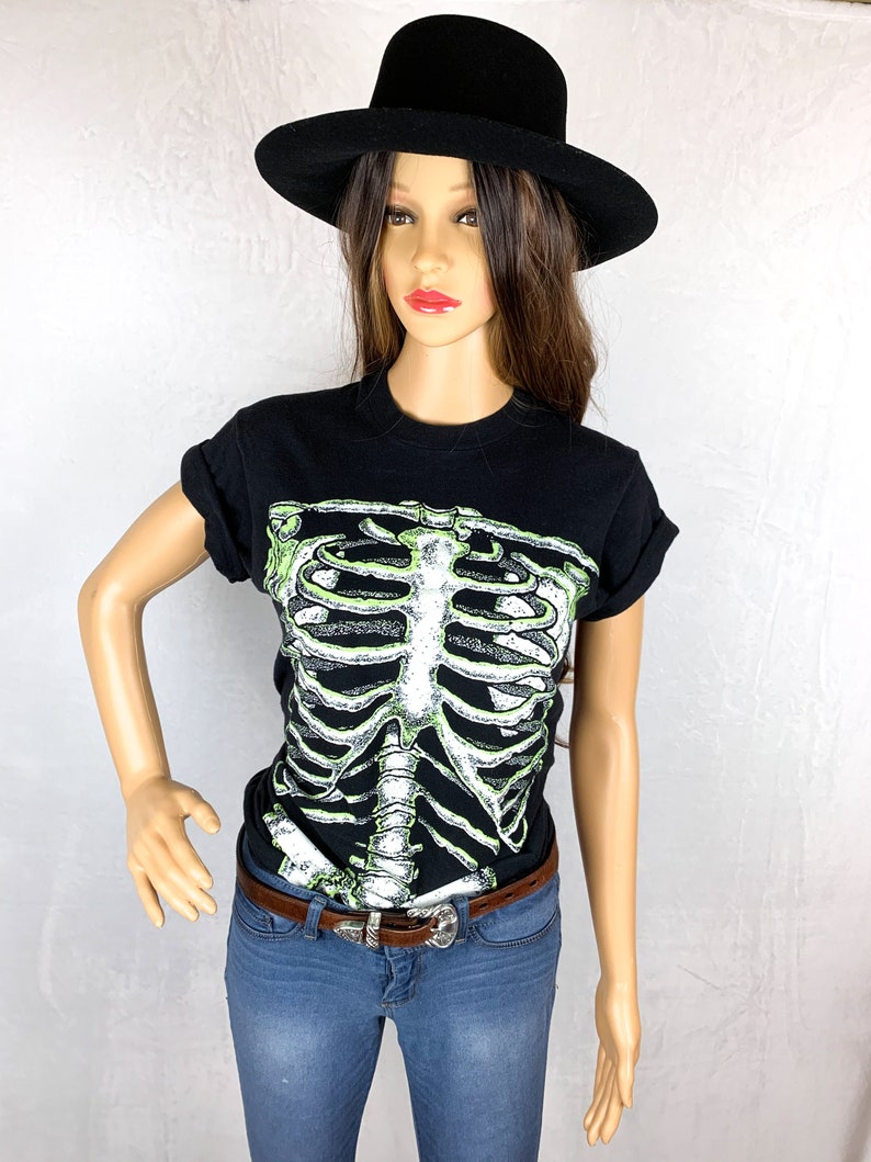 Op de afbeelding: Zwart T-shirt met een groene skeletprint. Het shirt heeft een ronde hals en korte mouwen. Het model draagt een zwarte hoed en een blauwe jeans.