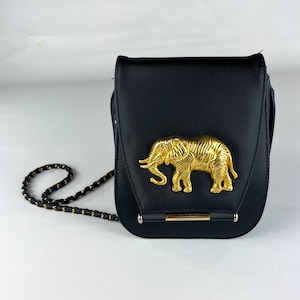 May include: Black crossbody bag with a gold elephant emblem. The bag has a textured surface and a gold chain strap. The elephant is detailed with intricate carvings. The bag has a rectangular shape with rounded edges.