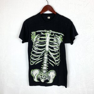 Op de afbeelding: Zwart T-shirt met een groene glow-in-the-dark skeletprint. Het skelet is een afbeelding van het hele lichaam, inclusief de ribbenkast, de wervelkolom en het bekken.