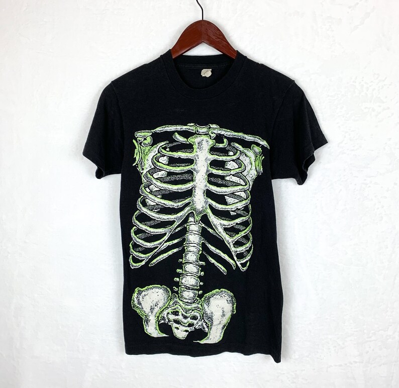 Op de afbeelding: Zwart T-shirt met een groene glow-in-the-dark skeletprint. Het skelet is een afbeelding van het hele lichaam, inclusief de ribbenkast, de wervelkolom en het bekken.