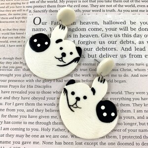 May include: A pair of white ceramic earrings shaped like a bear with black button eyes and a black nose. The earrings are hanging from a white post.