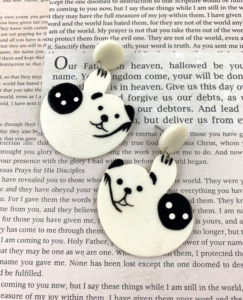 May include: A pair of white ceramic earrings shaped like a bear with black button eyes and a black nose. The earrings are hanging from a white post.