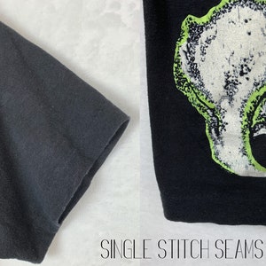 Op de afbeelding: Close-up van de zwarte stoffen mouw van een T-shirt met enkele steeknaden. De andere kant van de afbeelding toont een zwart T-shirt met een groene en witte grafische print van een schedel.