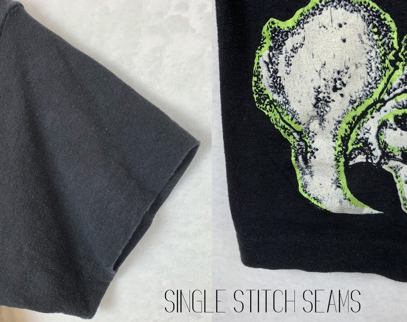 Op de afbeelding: Close-up van de zwarte stoffen mouw van een T-shirt met enkele steeknaden. De andere kant van de afbeelding toont een zwart T-shirt met een groene en witte grafische print van een schedel.