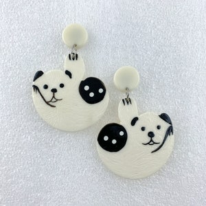 May include: A pair of white dangle earrings with a black and white panda bear design. The panda bears are holding a branch in their paws and have black eyes and noses.