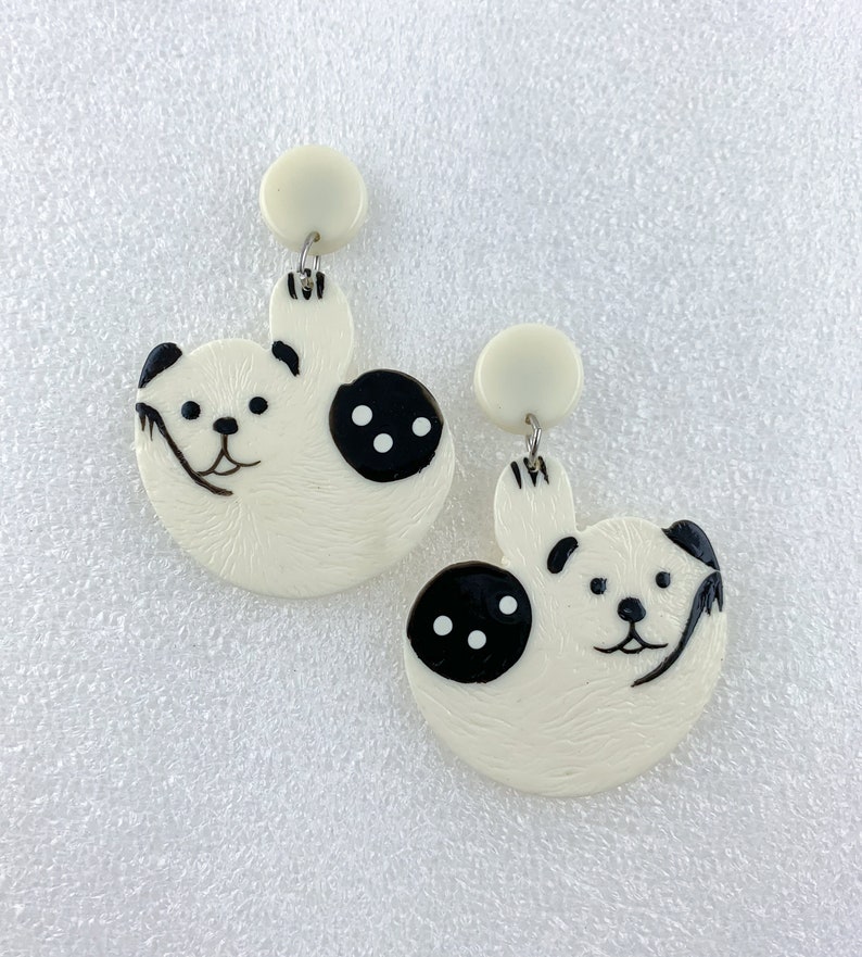 May include: A pair of white dangle earrings with a black and white panda bear design. The panda bears are holding a branch in their paws and have black eyes and noses.