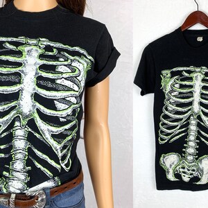 Op de afbeelding: Zwart T-shirt met een groene glow-in-the-dark skeletprint. De skeletprint omvat de ribbenkast, de wervelkolom en het bekken.