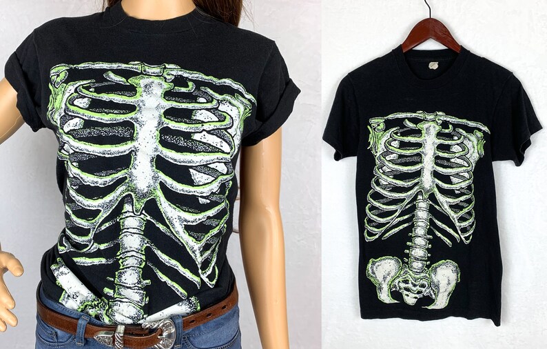 Op de afbeelding: Zwart T-shirt met een groene glow-in-the-dark skeletprint. De skeletprint omvat de ribbenkast, de wervelkolom en het bekken.