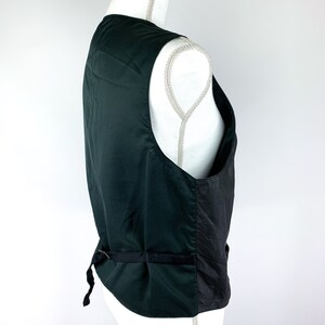 May include: A dark green waistcoat with a black satin back panel and a black adjustable strap. The waistcoat has a classic design and is made of a smooth fabric.