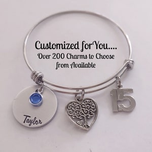 May include: Silver charm bracelet with a blue crystal charm, a heart charm with a tree design, and a charm with the number 15. The bracelet has a tag that says "Customized for You... Over 200 Charms to Choose from Available" and another tag that says "Taylor".