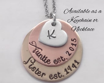 Puede incluir: Un collar de cadena plateada con un dije de capas que presenta las palabras "Auntie est. 2015" y "Sister est. 1941" sobre un fondo de cobre y oro. Un dije de corazón plateado con la letra "K" está unido a la parte superior del dije.