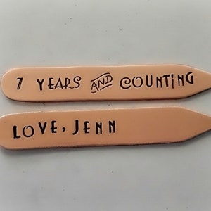 以下が含まれることがあります： 先が尖った銅色の金属製のタグが2つ。上のタグには「7 Years and Counting」と書いてあります。下のタグには「Love, Jenn」と書いてあります。