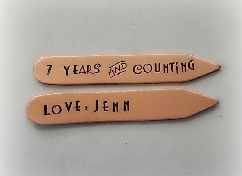 以下が含まれることがあります： 先が尖った銅色の金属製のタグが2つ。上のタグには「7 Years and Counting」と書いてあります。下のタグには「Love, Jenn」と書いてあります。