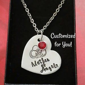 May include: Silver heart-shaped pendant necklace with a red gemstone and the inscription "Mother of Angels". The necklace is in a black gift box.