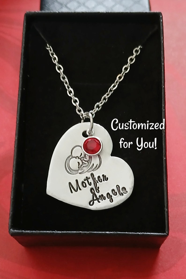 May include: Silver heart-shaped pendant necklace with a red gemstone and the inscription "Mother of Angels". The necklace is in a black gift box.