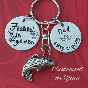 May include: Silver keychain with a fish charm, two round tags with the text "Fishin' in Heaven" and "Dad 1955 ♥ 2025", and a small angel wing design.