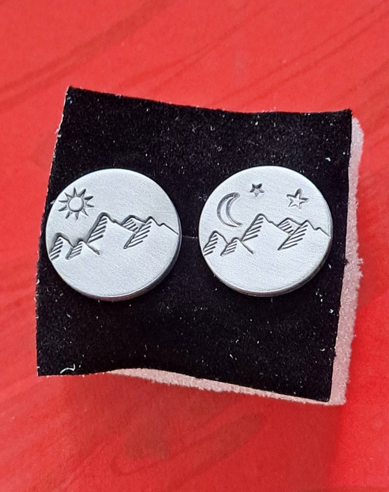 Puede incluir: Un par de pendientes de plata con un dise&ntilde;o de cordillera. Un pendiente tiene un sol y el otro tiene una luna creciente y dos estrellas.