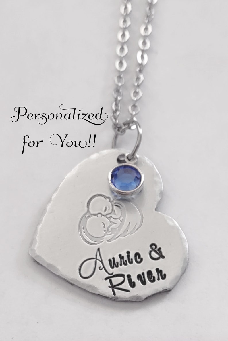 May include: A silver heart-shaped pendant necklace with a blue crystal charm. The pendant is engraved with the names "Auric & River" and a design of two babies sleeping.