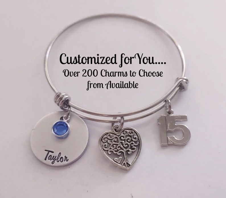 May include: Silver charm bracelet with a blue crystal charm, a heart charm with a tree design, and a charm with the number 15. The bracelet has a tag that says "Customized for You... Over 200 Charms to Choose from Available" and another tag that says "Taylor".