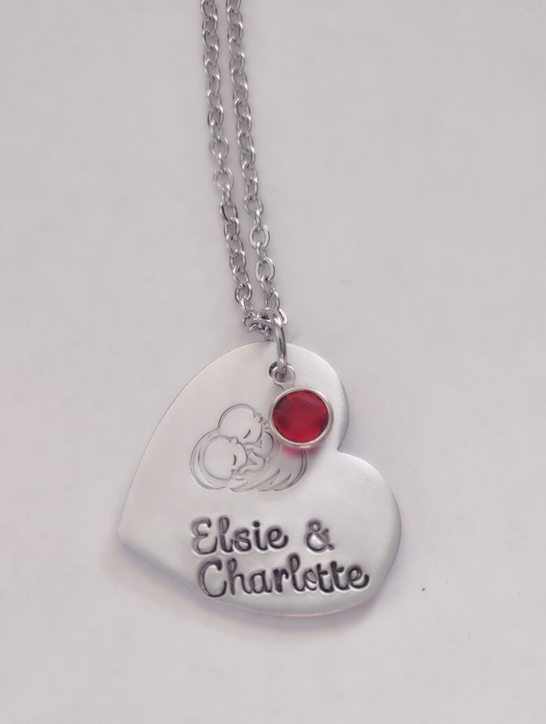 May include: A silver heart-shaped necklace with a red rhinestone charm. The pendant has an engraved image of two babies sleeping and the text "Elsie & Charlotte".