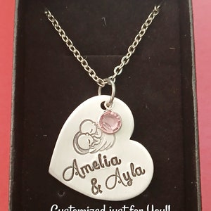 May include: Silver heart-shaped necklace with a pink crystal charm. The pendant features an engraved image of two babies and the names "Amelia & Ayla". The necklace is displayed in a black box with the text "Customized just for you!!"