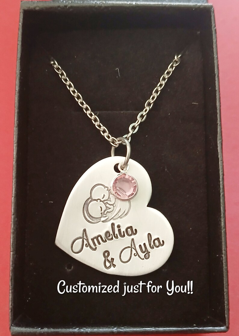 May include: Silver heart-shaped necklace with a pink crystal charm. The pendant features an engraved image of two babies and the names "Amelia & Ayla". The necklace is displayed in a black box with the text "Customized just for you!!"