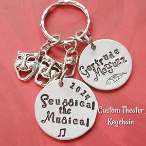 Op de afbeelding: Zilveren sleutelhanger met een ronde hanger waarop "Seussical the Musical" staat met een muzieknotensymbool en het jaar 2024. Een andere ronde hanger heeft de tekst "Gertrude McFuzz" met een verenontwerp. De sleutelhanger heeft ook twee kleine zilveren hangers in de vorm van theatermaskers. De tekst "Custom Theater Keychain" is onder de hangers gedrukt.