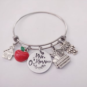 Könnte beinhalten: Silbernes Armband mit Charms, darunter ein Schulhaus, ein Apfel, Bücher und eine Eule. Das Armband hat einen runden Anhänger mit dem Text "Mrs. O'Morrow".