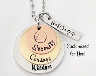 Może przedstawiać: Srebrny naszyjnik z wielopoziomowym wisiorkiem przedstawiającym symbol trójkąta, słowa "Serenity", "Courage" i "Wisdom" oraz srebrną etykietę z datą "8.10.22".
