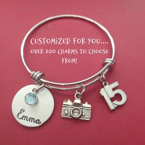 May include: Silver charm bracelet with a camera charm, a charm with the number 15, and a personalized charm with the name "Emma" and a light blue gemstone. The bracelet is on a red background.  "CUSTOMIZED FOR YOU... OVER 200 CHARMS TO CHOOSE FROM!"