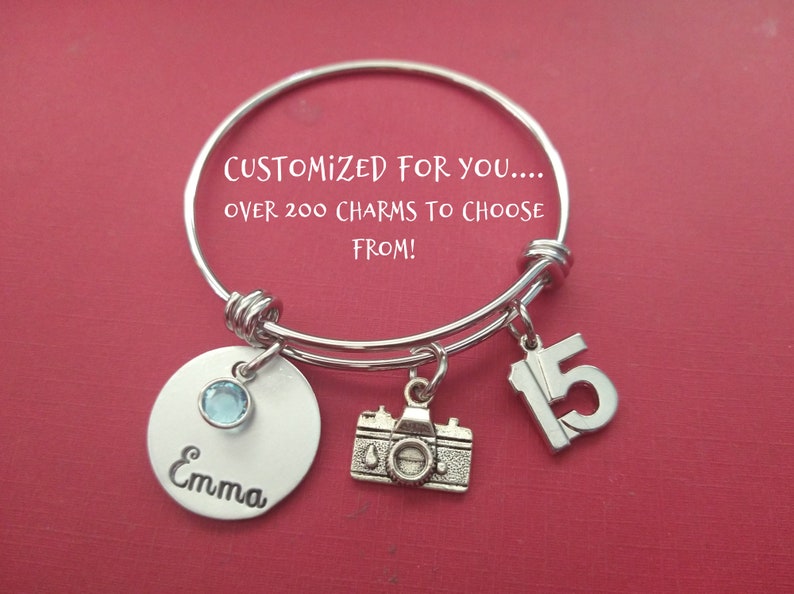 May include: Silver charm bracelet with a camera charm, a charm with the number 15, and a personalized charm with the name "Emma" and a light blue gemstone. The bracelet is on a red background.  "CUSTOMIZED FOR YOU... OVER 200 CHARMS TO CHOOSE FROM!"