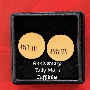 Puede incluir: Un par de gemelos de color dorado en una caja negra. Los gemelos son redondos y tienen marcas de conteo grabadas en ellos. El texto "Anniversary Tally Mark Cufflinks" está impreso debajo de los gemelos. El texto "Available in Bronze, Copper or Aluminum" está impreso debajo de la caja.