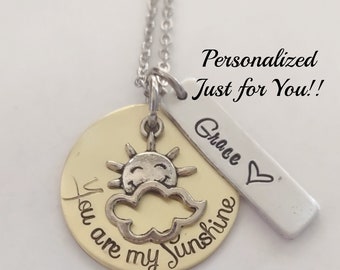 Op de afbeelding: Gouden ketting met een ronde hanger waarop "You are my sunshine" staat met een zilveren zon- en wolkbedel. Een zilveren tag met de naam "Grace" is aan de ketting bevestigd.