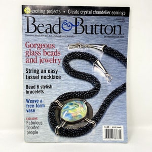 Puede incluir: Una portada de revista para Bead & Button, que presenta un collar de cuentas negras con un cierre plateado y un colgante de cuentas de vidrio de colores. El texto de la portada dice "Preciosas cuentas de vidrio y joyas".