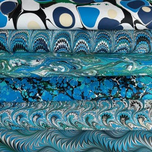 May include: A selection of marbled paper sheets in shades of blue, teal, and white. The patterns vary, including swirls and organic shapes. The paper has a handmade appearance, with a unique artistic quality. The sheets are stacked together.