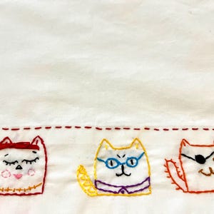 May include: Three embroidered cats on a white fabric background. The cat on the left is wearing a red headband and has closed eyes. The cat in the middle is wearing glasses and has a yellow tail. The cat on the right is wearing an eye patch and has a brown tail.