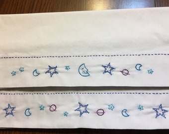 Puede incluir: Tela blanca con diseños celestes bordados. El bordado presenta estrellas, lunas y planetas azules. Una línea azul discontinua recorre la parte superior e inferior del patrón bordado. El diseño se repite dos veces.